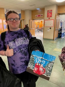 A Ridge View Community School student holds up a painting of a red mug framed by snowy pine trees on a bright blue landscape while giving the camera a thumbs up. 