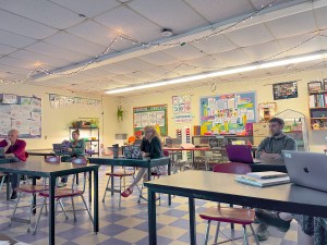 Teachers sit at rectangular tables listening to a woman in the center of the room speak.