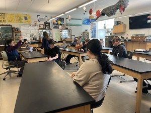 Mt. Blue BARR Students sit in a science classroom at long black lab desks, facing the front of the room where a teacher sits in front of a laptop talking to them. 