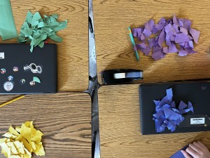 Torn piles of paper on wooden school desk tops with four piles of paper, one green, one purple, one blue and one yellow, with a black tape dispenser.