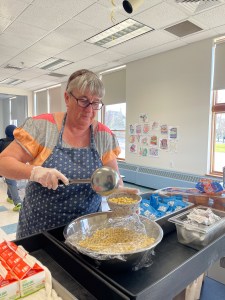 A woman in a hair net and apron scoops kicks cereal into a white bowl. 