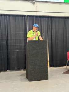 A student in a bright green shirt and blue hat stands at a black podium giving a speech.