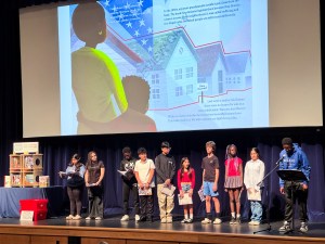 Students line a stage with books in their hands. On the screen is an illustration of a son hugging his mother around the waist, looking out at houses. To the far left of the state is a book shelf filled with books.
