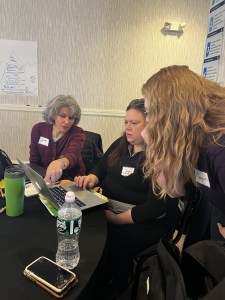 Three women sit at the end of a table. The woman on the far left points at the computer screen of the woman in the middle, and the woman on the right leans over to look at the screen.