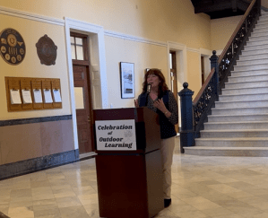 Commissioner Pender Mekin stands at a wooden podium with a white sign that says “Celebration of Outdoor Learning” in green. The Commissioner is speaking and holding her hands up in emphasis.