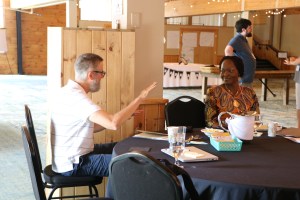 A man and a woman sit at a table. The man is speaking, illustrating his point with a hand held in the air. 