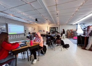 Students sit at desks grouped in fours facing each other. A teacher, Melanie Stevens, walks behind them reading an I-Time story off a piece of paper. In the fair right, Matthew Foster observes the BARR class.